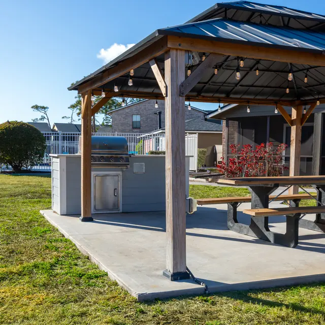 A well-maintained outdoor pavilion featuring a covered picnic area with wooden tables, surrounded by lush grass and decorative shrubs, near a swimming pool area.