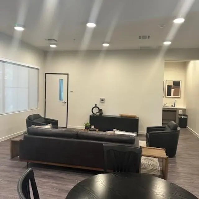 Modern Living Room Interior A modern living room with a sectional sofa, coffee table, and dining table. White walls and recessed lighting create a bright atmosphere. A door and a window are visible on the left side of the image, and a kitchen area is seen in the background.