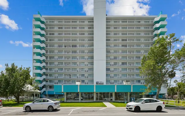 A modern hotel building with a turquoise and white exterior, featuring multiple stories and balconies. The building is set against a bright blue sky with some clouds. In front of the hotel, there are cars parked on the street and trees lining the area.