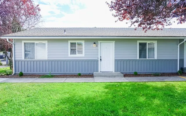 Gray House Front View A single-story gray house with a front lawn, featuring a white door and several windows. The lawn is green and well-maintained with small shrubs along the pathway.