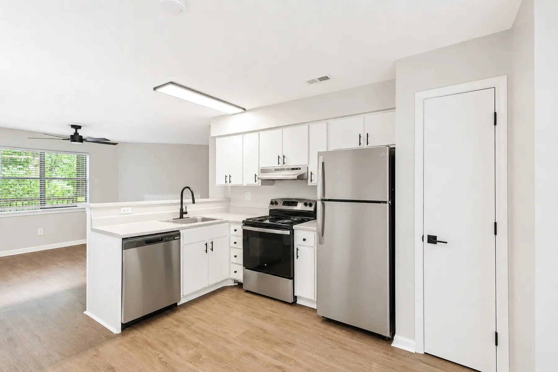A modern kitchen with stainless steel appliances, including a refrigerator and stovetop. The cabinets are white, and the countertop is light, with a sink and faucet. The flooring is wood-like, and there are windows that provide natural light into the space.