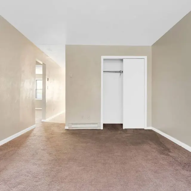 An empty room with brown carpet and light beige walls. A closet with white doors is visible on the right, and a hallway can be seen in the background leading to a well-lit area.