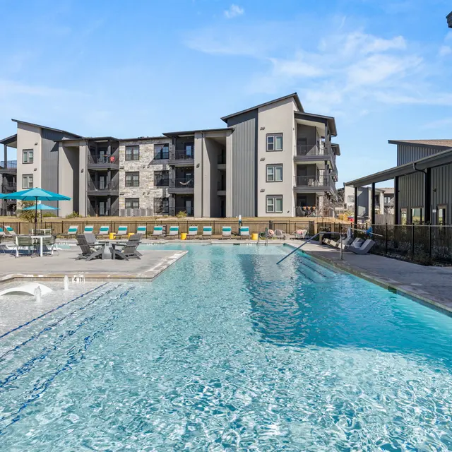 A modern apartment complex with a large swimming pool in the foreground, featuring lounge chairs and umbrellas. The pool is surrounded by a clean deck area and beyond it, there are multiple apartment buildings.
