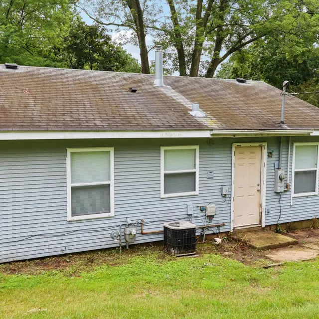 A single-story blue house with a plain exterior, featuring three windows and a central door. The lawn is green and well-kept, and there is a black air conditioning unit on the side of the house.