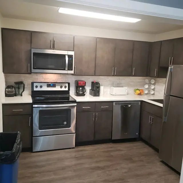 Modern kitchen with stainless steel appliances including an oven, microwave, and refrigerator, dark cabinetry, and wood-like flooring.