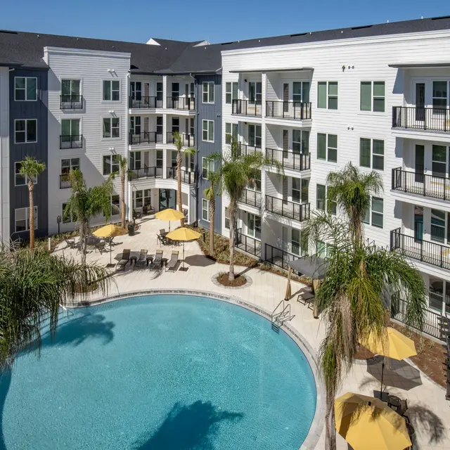An aerial view of a modern apartment complex featuring a circular pool surrounded by palm trees and yellow umbrellas. The building has a mix of white and gray exteriors with multiple balconies.
