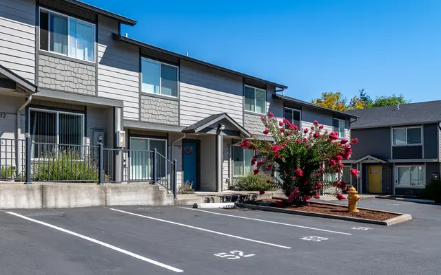 Exterior view of a residential apartment complex, featuring two-story buildings with a parking area in front, colorful flowers, and clear blue skies.