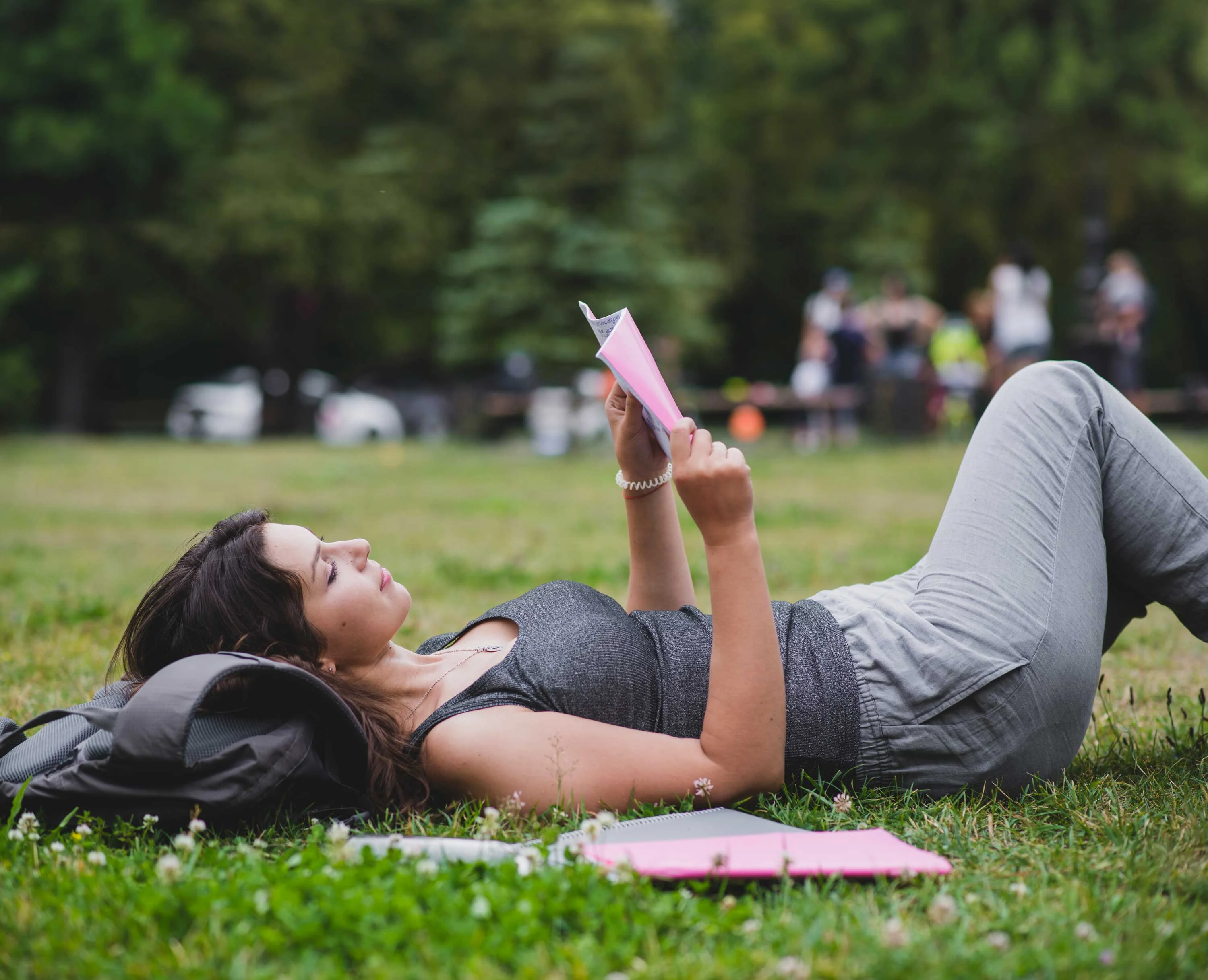 A woman lying on the grass in a park, reading a pink booklet while surrounded by greenery.