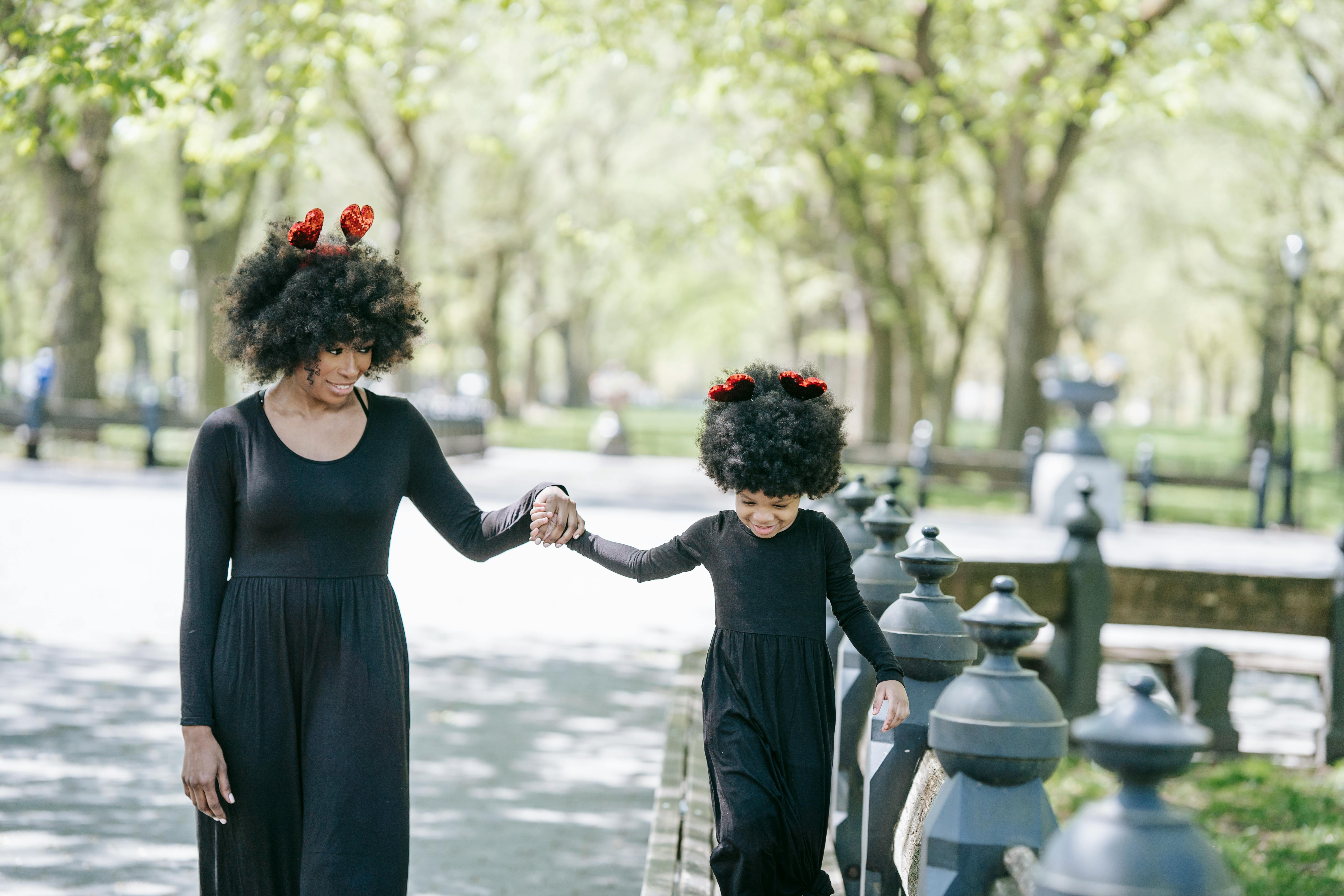 Mother and Child in a Park A woman and a young girl walking hand in hand in a park, both wearing matching black dresses and red hair accessories with a sunny background.