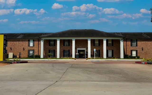 A brick apartment building with white columns and a flat roof, surrounded by green landscaping under a partly cloudy sky.