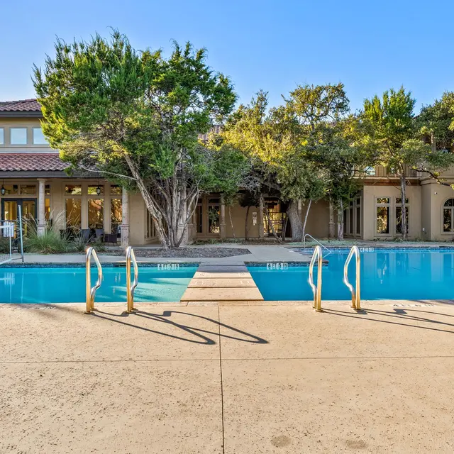 A serene view of a swimming pool with a tan concrete deck, surrounded by lush greenery and a modern building in the background.