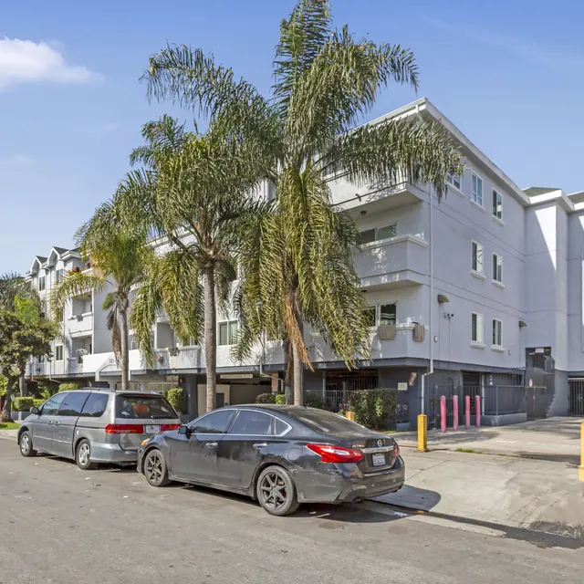 A street view of a white apartment building lined with palm trees. Several parked cars are visible in front of the building, alongside a road with sparse traffic. The sky is clear and blue.
