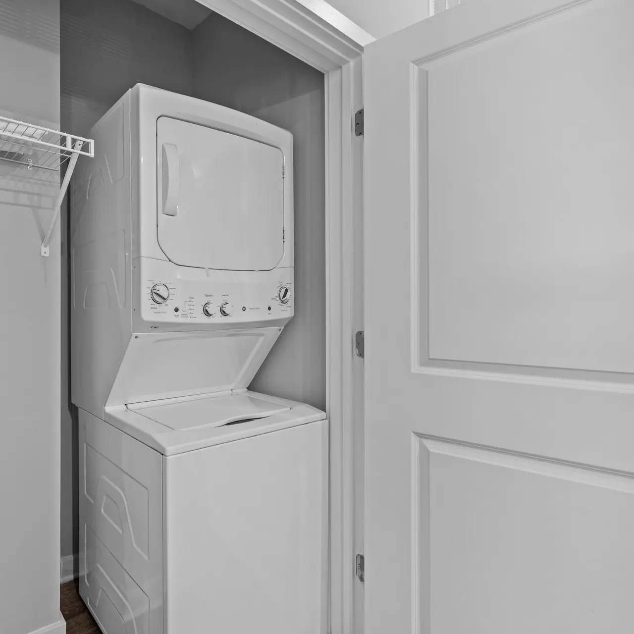 A compact laundry area featuring a stacked washer and dryer inside a closet. The walls are painted light gray, and there is a wire shelf above the appliances.
