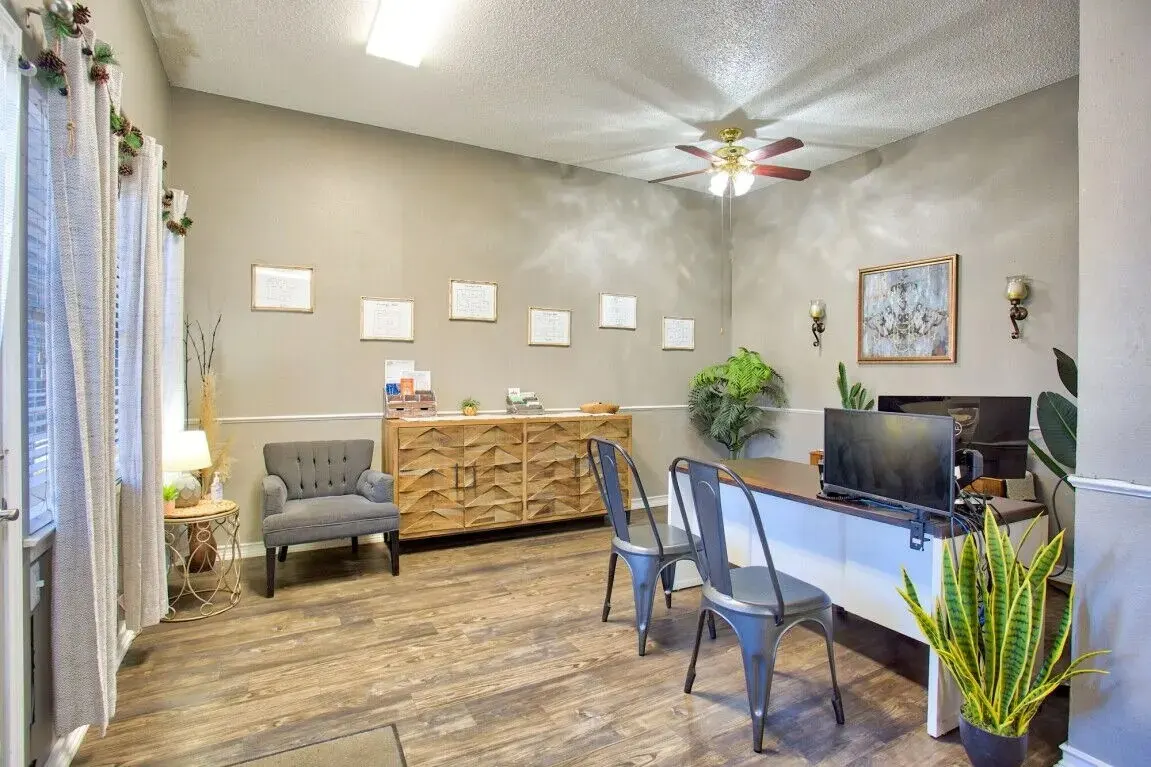 A modern office interior featuring a light gray wall, wooden furniture, and two metal chairs. There is a comfortable gray armchair in the corner, a large wooden cabinet, and greenery decor including a potted plant. The room is well-lit with a ceiling fan and wall sconces.