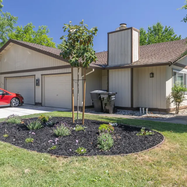 A single-story house with beige siding, a brown roof, and a red car parked in the driveway. There are landscaping features including a circular garden bed filled with small plants and mulch in the foreground.