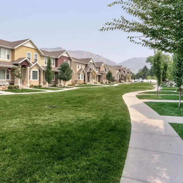 A serene suburban neighborhood featuring neatly arranged houses along a wide, grassy area, with sidewalks lined by young trees, and mountains visible in the background under a clear sky.