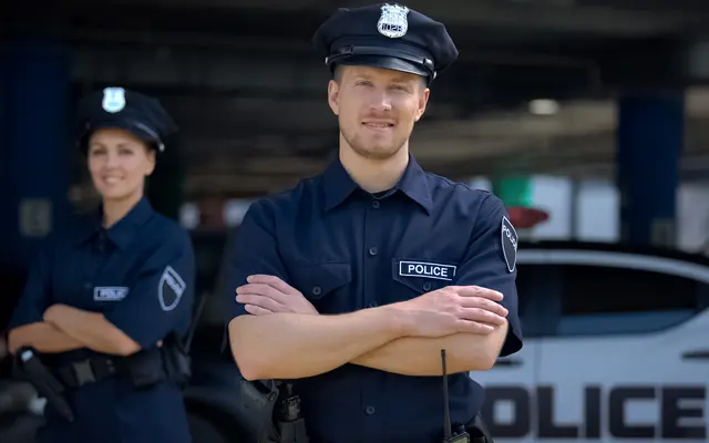 Oaks of Kingwood Two police officers in uniform stand with arms crossed in front of a police car, with one officer in focus and the other blurred.
