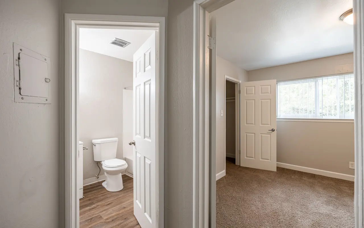 Interior view showing a bathroom door on the left and a bedroom door on the right, with a window in the bedroom and features including carpeting and neutral-colored walls.