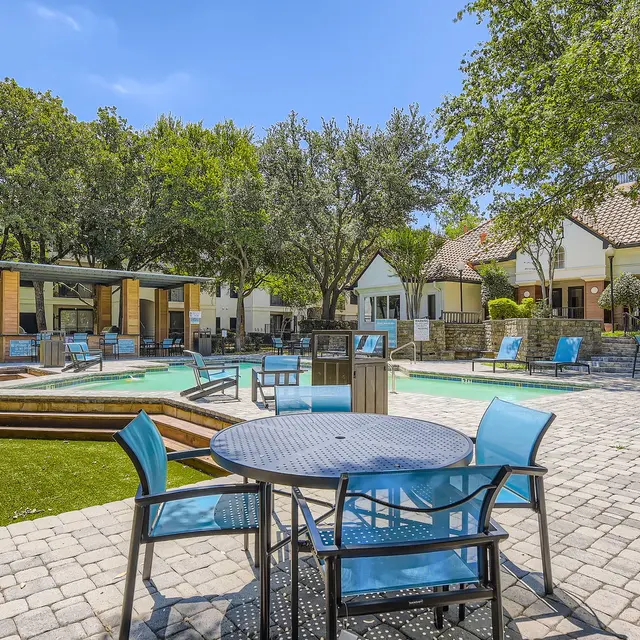 A beautifully designed outdoor pool area featuring lounge chairs, umbrellas, and tables with chairs. There are trees and buildings in the background, with a brick walkway leading to the pool.