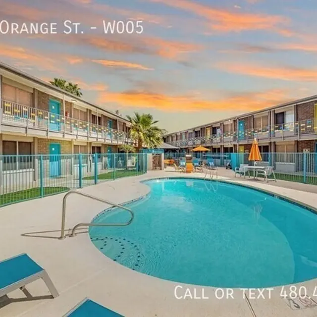 A swimming pool surrounded by a courtyard with palm trees and lounge chairs, and buildings with balconies in the background, under a colorful sunset sky.