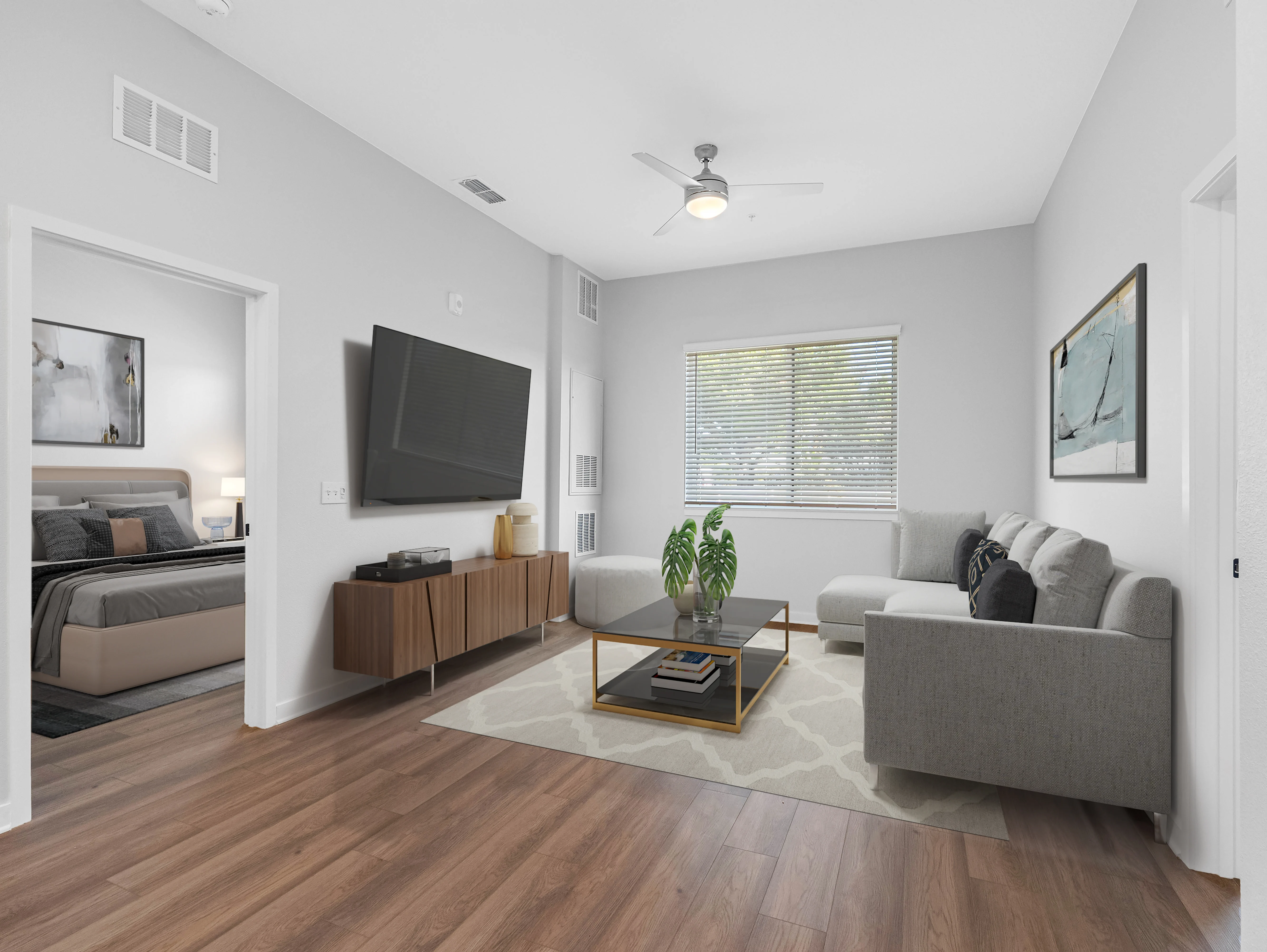 A modern living room featuring a comfortable gray sectional sofa, a round coffee table, and a large wall-mounted television. Natural light pours in through the window, illuminating the wood floor and light-colored walls.