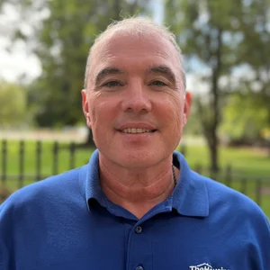 A man smiling outdoors in a blue shirt, with green trees in the background.