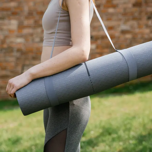 A person carrying a rolled yoga mat over their shoulder in an outdoor setting with green grass and a blurred brick wall in the background.