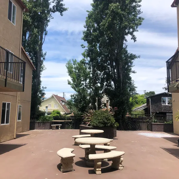 A spacious courtyard surrounded by two buildings, featuring a circular arrangement of stone benches and a tree in the center. The sky is partly cloudy and there are green trees on both sides.