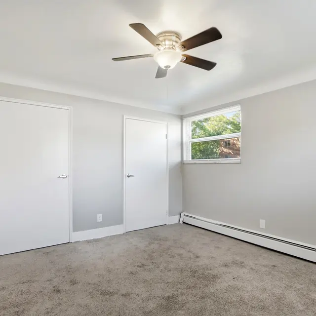 A spacious empty bedroom featuring light gray walls and carpet, a ceiling fan, and two closed doors on the left side, with a window letting in natural light.