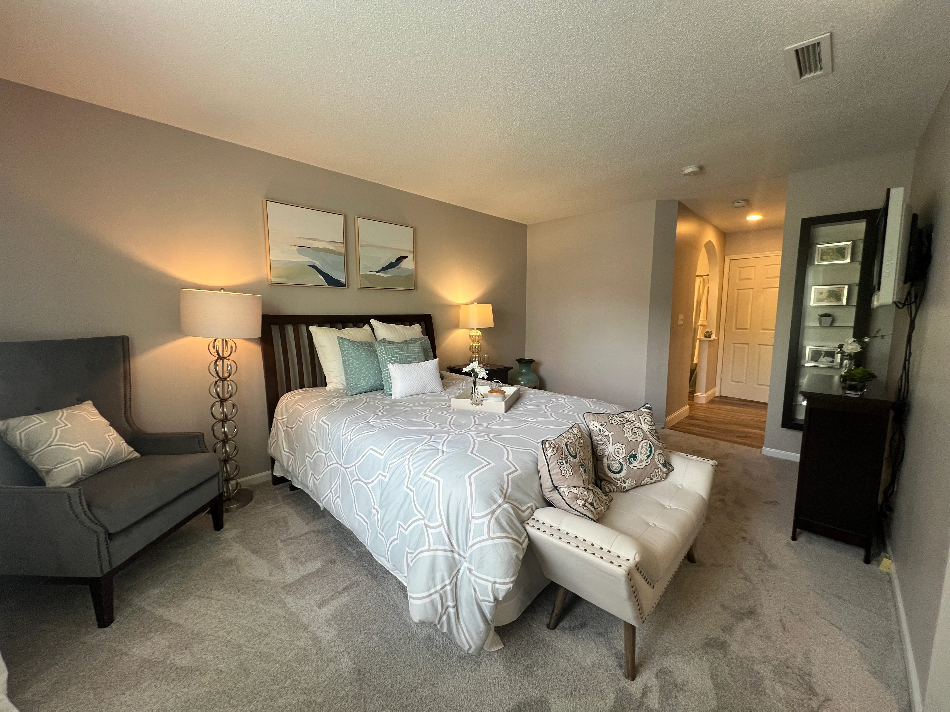 A cozy bedroom featuring a double bed with light-colored bedding, two bedside lamps, a stylish armchair, and a view of an adjacent hallway.