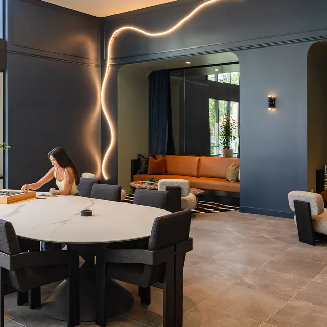 A modern lounge area featuring a large round table with two people playing a game, surrounded by stylish chairs and a cozy seating area with an orange couch and decorative elements.