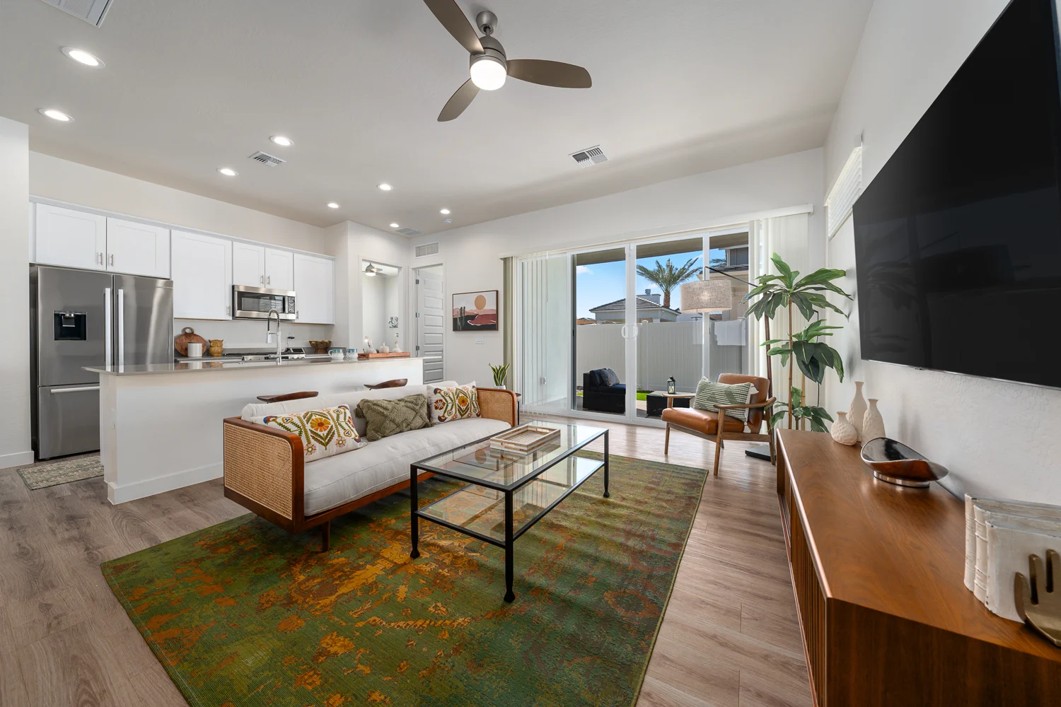Bright and Modern Living Room A modern living room featuring a light-colored sofa with decorative cushions, a glass coffee table, and a green area rug. The kitchen is visible in the background with white cabinets and stainless steel appliances. Large sliding doors lead outside, allowing natural light to fill the space. There's a plant in the corner and wooden furniture complementing the decor.
