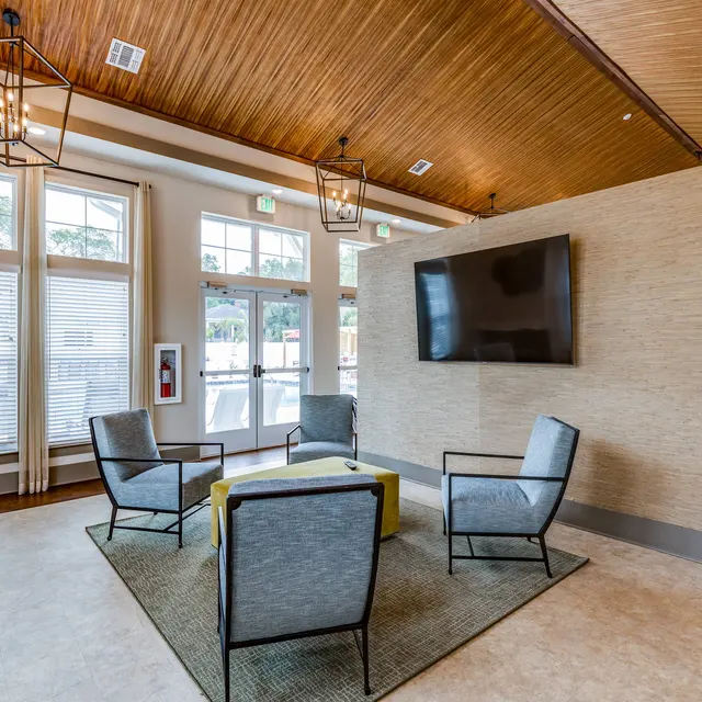 ShoreView A sitting area in a clubhouse with four chairs, a large ottoman, and TV on a partition wall near doors leading to a pool.