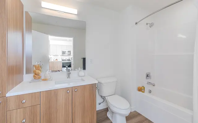 A modern bathroom featuring wooden cabinetry, a white countertop, a toilet, and a bathtub with a shower. A mirror reflects the kitchen area visible in the background.