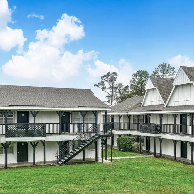 A view of a two-story apartment complex with white and gray buildings, featuring balconies and green grass in the foreground under a blue sky with clouds.