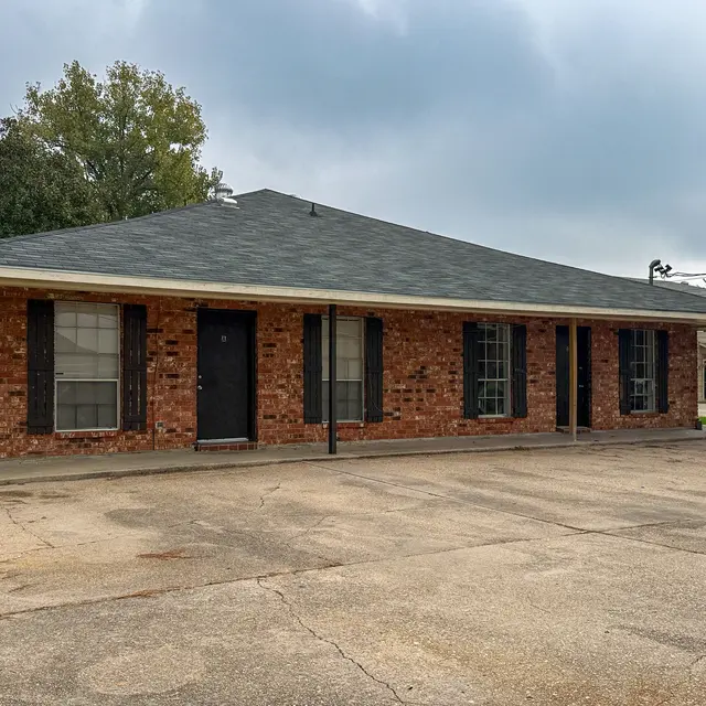A view of a single-story brick house with a gray roof, featuring black shutters and a front porch area. The driveway is made of concrete with some cracks visible. In the background, there are trees and another building.