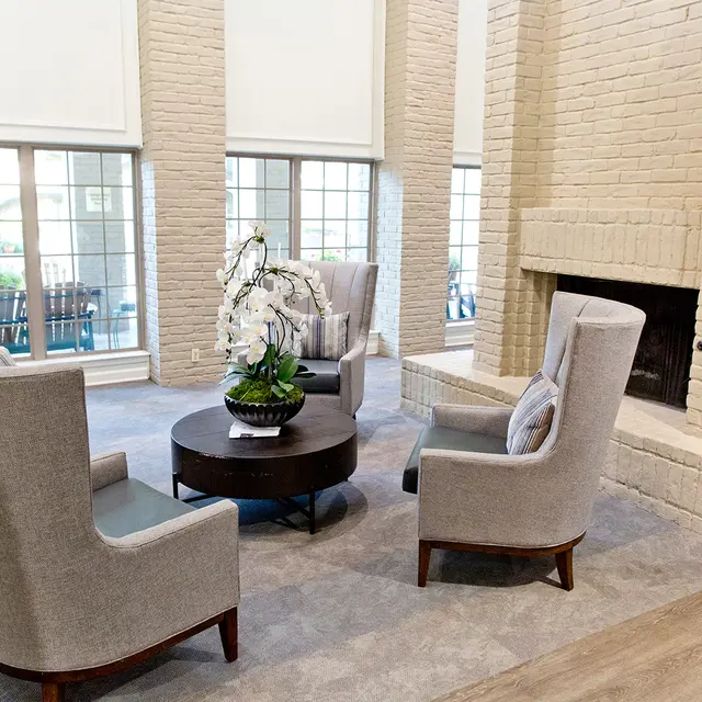 A modern living room with a cozy seating area featuring gray upholstered armchairs, a round coffee table with a plant centerpiece, and a fireplace against a brick wall.