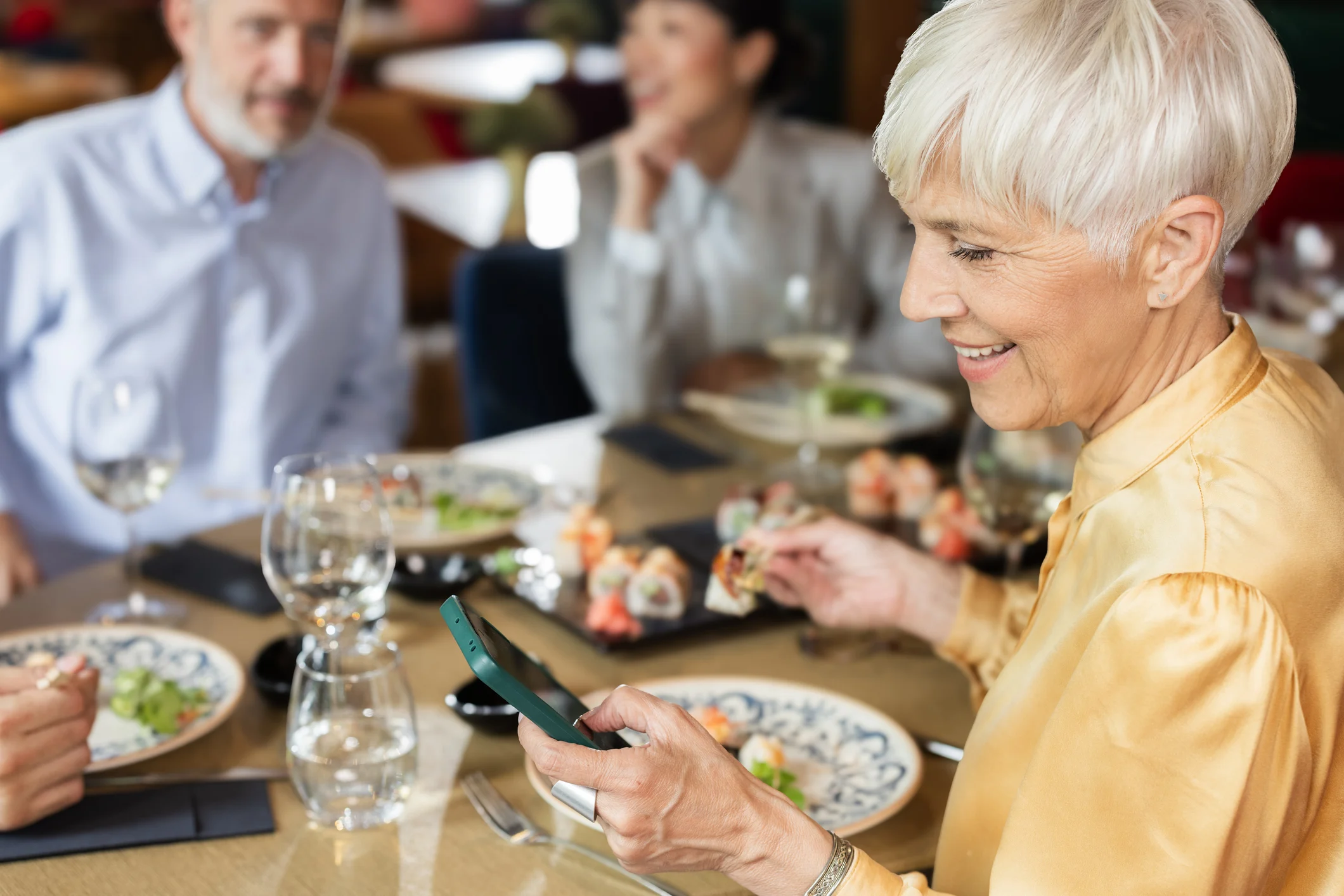 A group of friends enjoying a meal together in a restaurant, with a focus on a smiling older woman in a golden blouse holding a smartphone and a piece of sushi, while others are seated at the table.