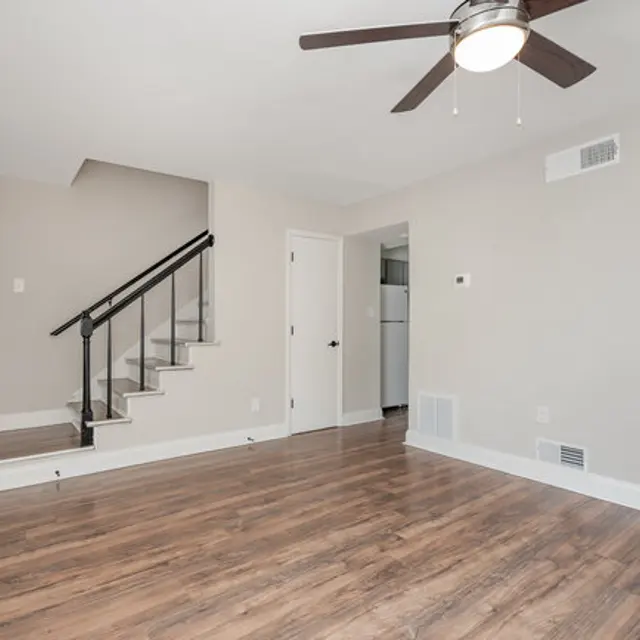 A spacious living room featuring wooden flooring, neutral-colored walls, and a ceiling fan. There is a staircase on the left leading to an upper level, with a door to the right leading to a kitchen area. Natural light illuminates the space.