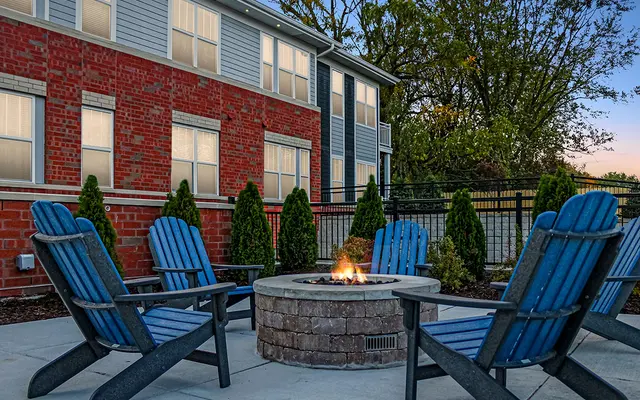 Outdoor seating area with blue Adirondack chairs around a fire pit near a red brick building at dusk.
