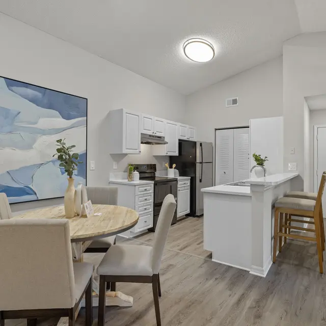 A modern kitchen with a dining area featuring a round table and beige upholstered chairs, a large blue abstract painting on the wall, white cabinets, and stainless steel appliances.