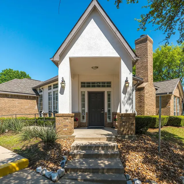 A welcoming community center with a peaked roof and stone accents. The entrance features a set of steps leading up to the front door, surrounded by neatly landscaped greenery. There are trees and open space visible in the background under a clear blue sky.