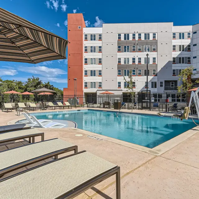 A modern apartment pool area with lounge chairs and umbrellas, featuring a swimming pool surrounded by greenery and a multi-story building in the background.