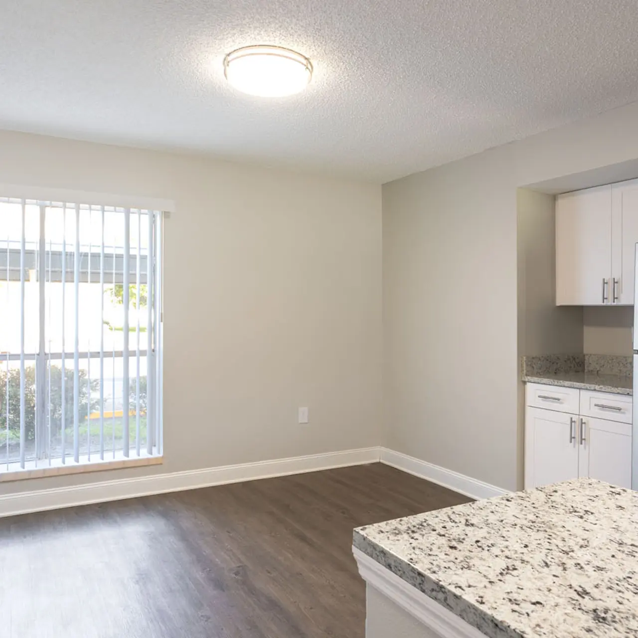 An interior view of a modern apartment featuring a spacious living area with a large window and blinds, light-colored walls, wood-style flooring, and a kitchen area with granite countertops and stainless steel appliances.