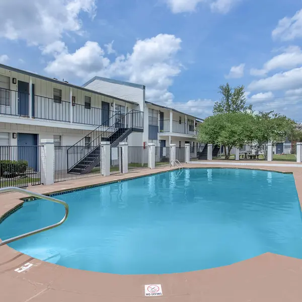 Swimming pool area surrounded by two-story apartment buildings, with clear skies and lush greenery.