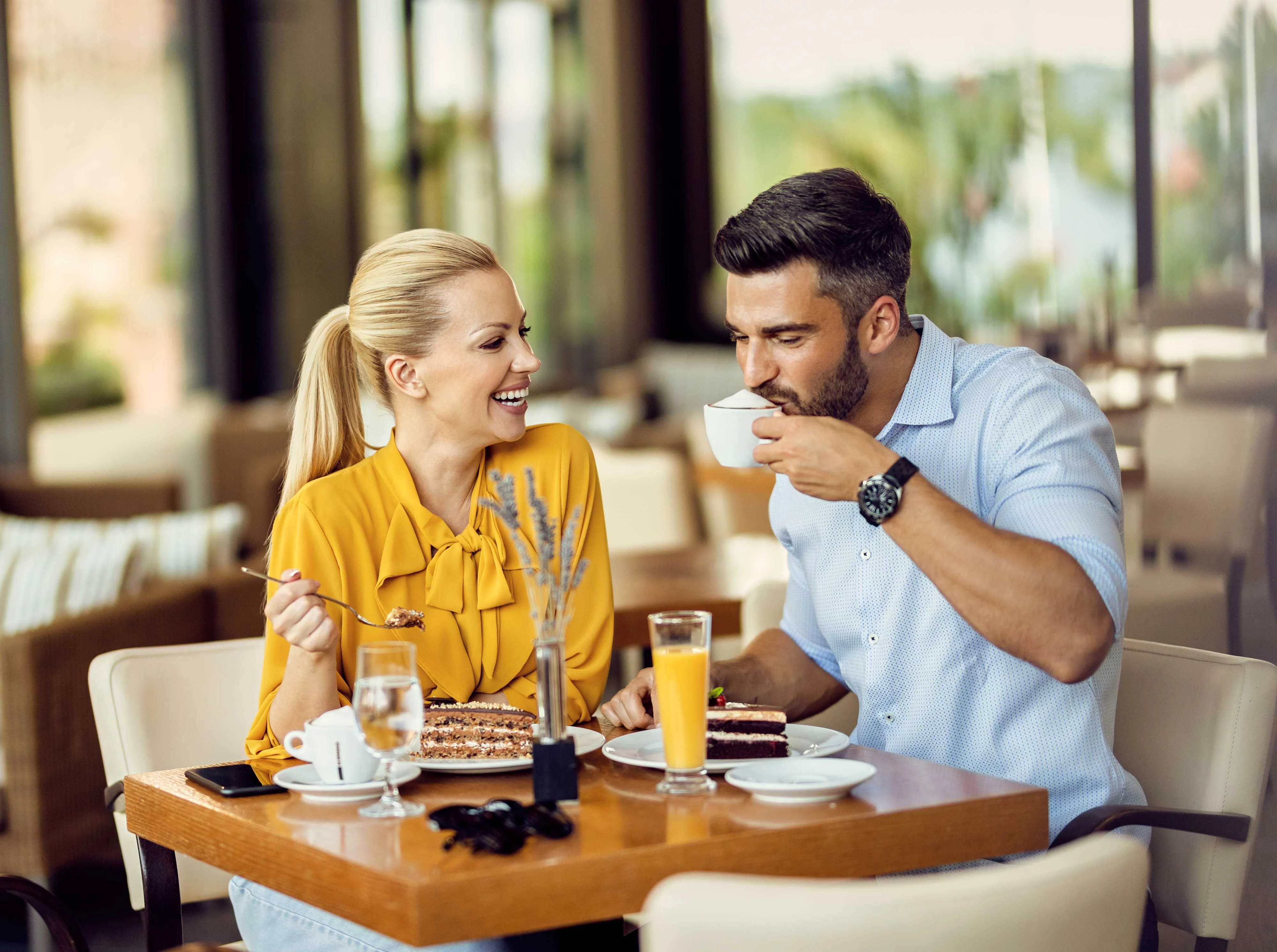 A smiling couple sitting at a table in a restaurant, enjoying dessert and drinks. The woman is wearing a yellow blouse and holding a fork, while the man is sipping from a cup.