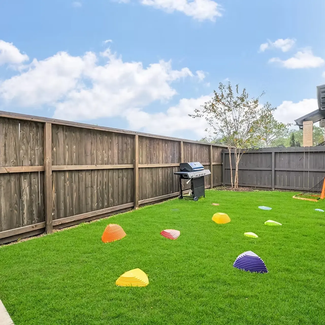 A backyard featuring a green lawn with colorful play cones scattered across it, a charcoal grill by the fence, and a tree in the background under a blue sky