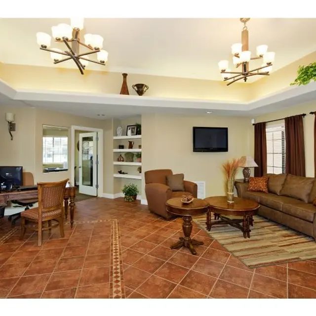 Comfortable Living Room Interior A cozy living room featuring tan walls, a brown sofa, a wooden coffee table, and a desk with a computer. There are decorative plants and large windows with dark curtains. The space has a chandelier and a mounted TV on the wall.