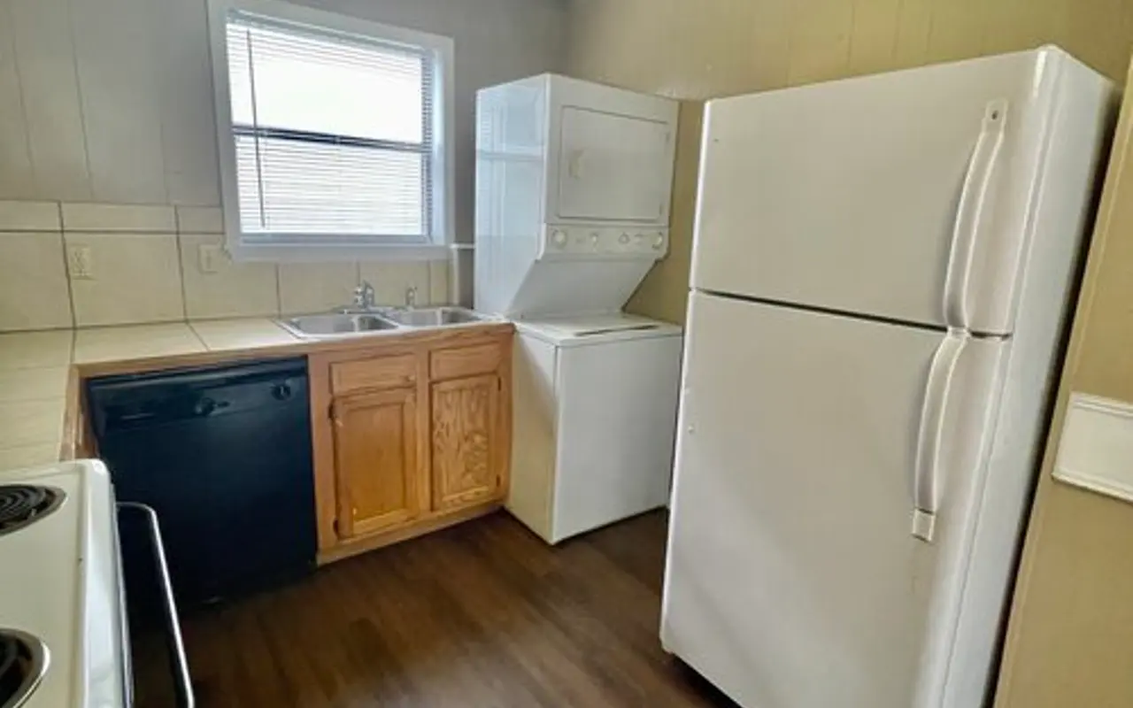 Compact Kitchen with Appliances A compact kitchen featuring basic appliances including a refrigerator, stove, and laundry area with a washer and dryer combo. The kitchen has wooden cabinets and a small window letting in natural light.