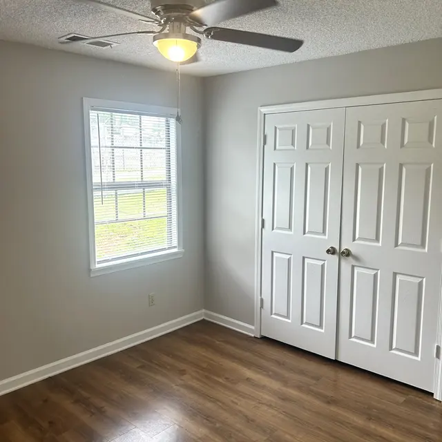 A bright, empty room featuring a ceiling fan and a window. There are white double doors on one wall and light wooden flooring throughout.